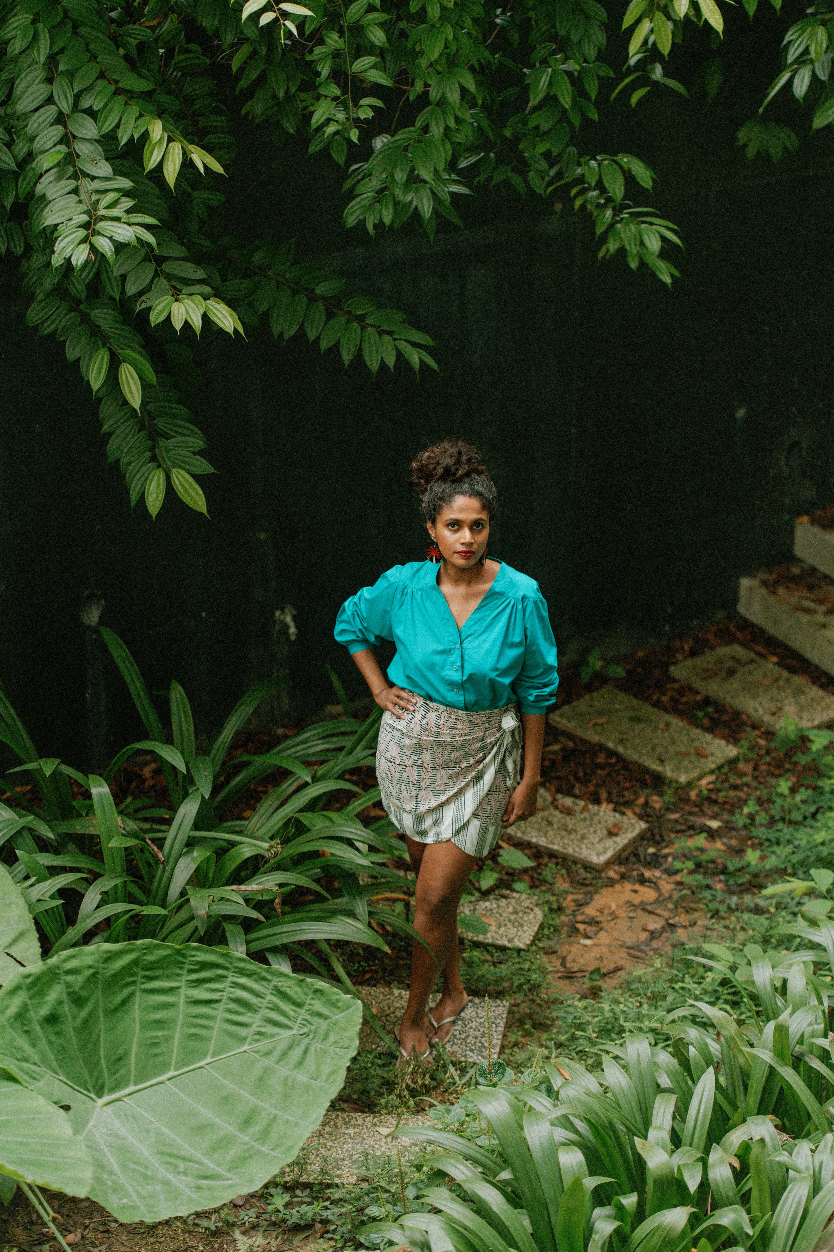 Woman in a blue shirt and patterned skirt standing on a stone path surrounded by greenery.